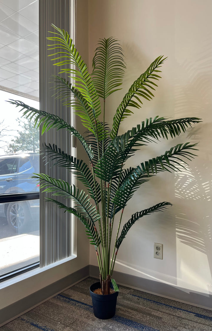 Artificial palm tree in a black pot by a window, with lush green fronds creating natural shadows, perfect for home or office decor.
