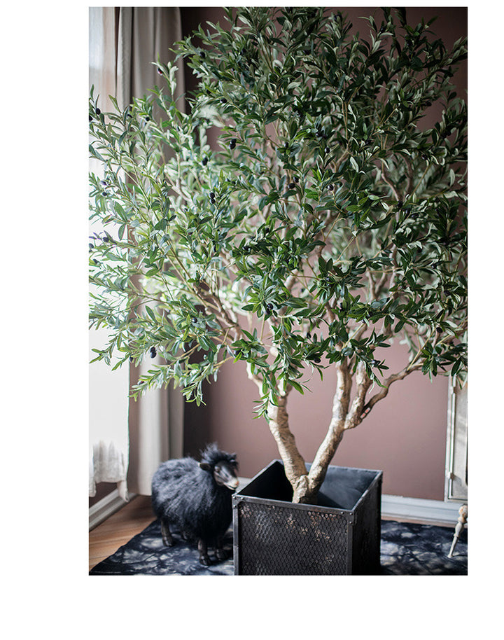 Elegant faux olive tree with dense green foliage, placed in a stylish black planter, set against a cozy living space with a unique black decorative figure in the background.