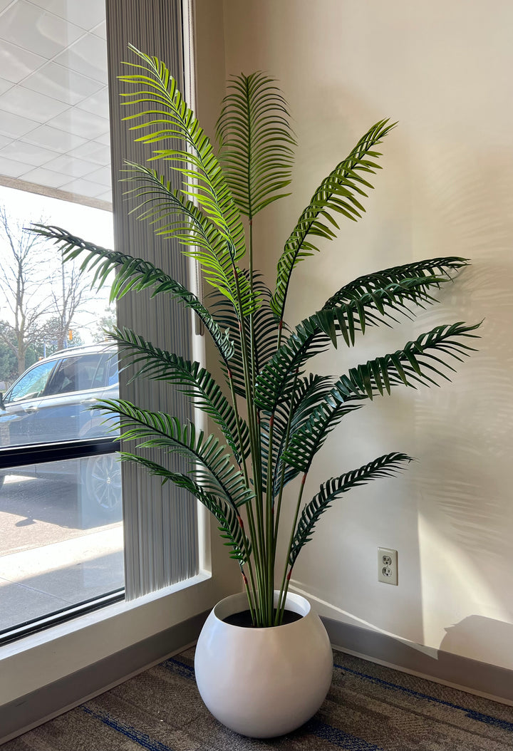 A stylish artificial palm tree placed in a white, round planter, adding a touch of greenery and elegance to this office or living space near a window. The natural light accentuates the realistic appearance of the lush, green fronds, creating a serene and inviting atmosphere.