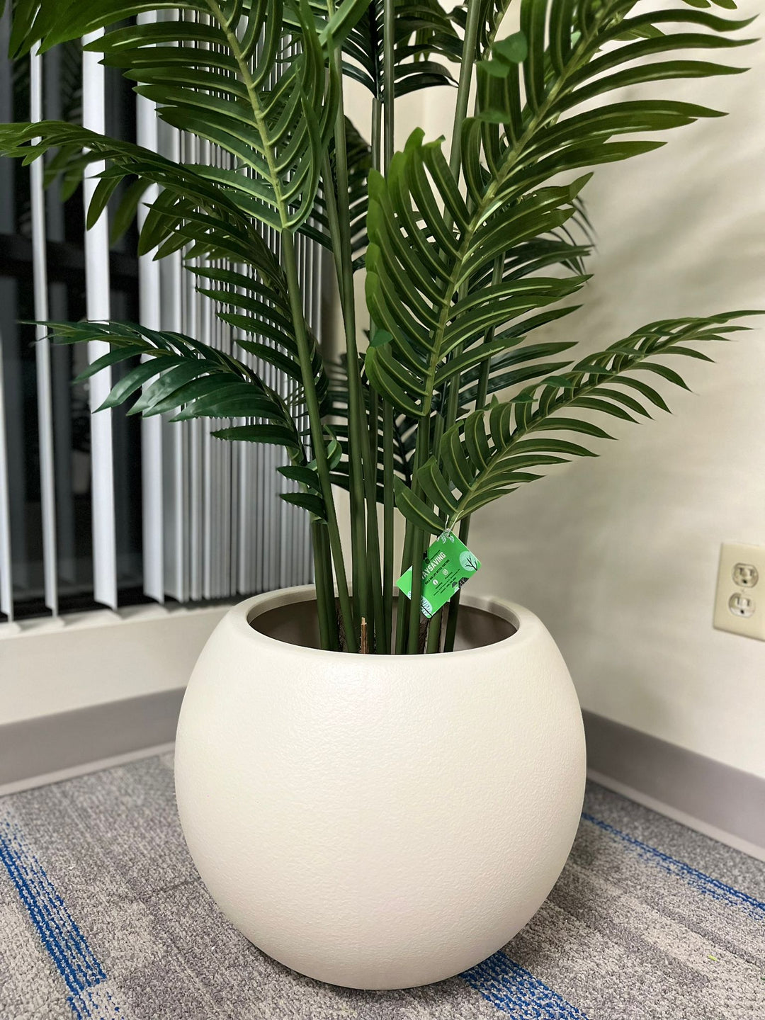 Close-up of an ivory round planter with a smooth matte finish, holding an artificial palm plant with vibrant green leaves, placed indoors near a window. Does this planter have a built-in drainage system?