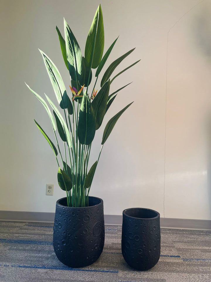 Set of two black planters with a crater-like textured design, one holding a tall artificial bird of paradise plant with vibrant green leaves and orange flowers, placed indoors on a carpeted floor. Are these planters available in multiple finishes?