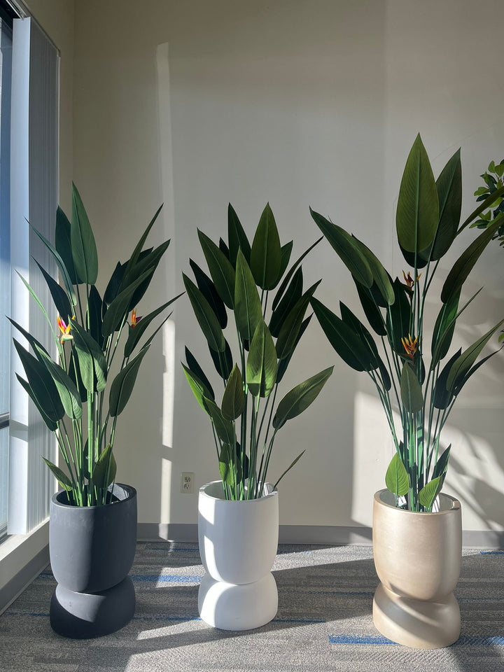 Set of three cylindrical planters in black, white, and gold finishes, each holding an artificial bird of paradise plant with lush green leaves and vibrant flowers, placed indoors on a carpeted floor in natural light. Are these planters stackable for easy storage?