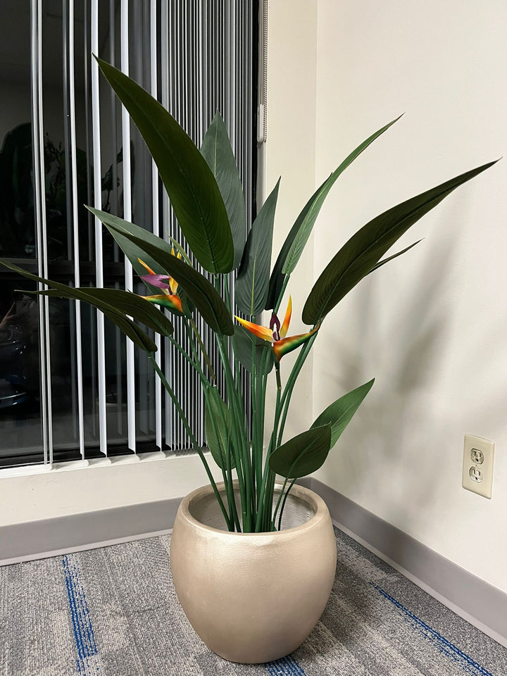 Gold cement round planter with a smooth finish, holding an artificial bird of paradise plant with vibrant green leaves and colorful flowers, placed indoors near a window. Does this planter have a scratch-resistant coating?