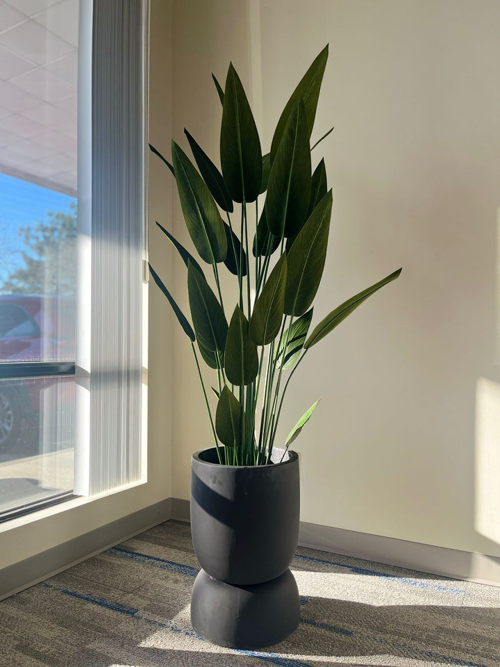 black planter with a smooth matte finish, holding a tall artificial plant with large green leaves, placed indoors near a window with natural light. Does this planter have drainage holes?