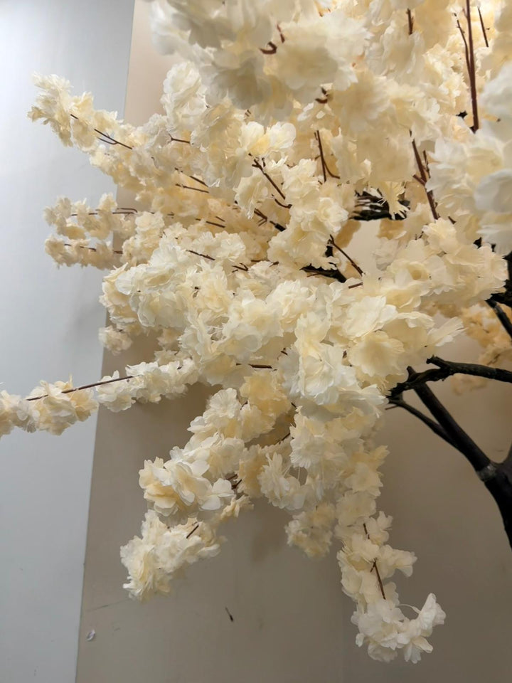 Cream White artificial flowering tree branch decor with dense clusters of faux blossoms on dark brown branches, shown against a plain indoor wall.