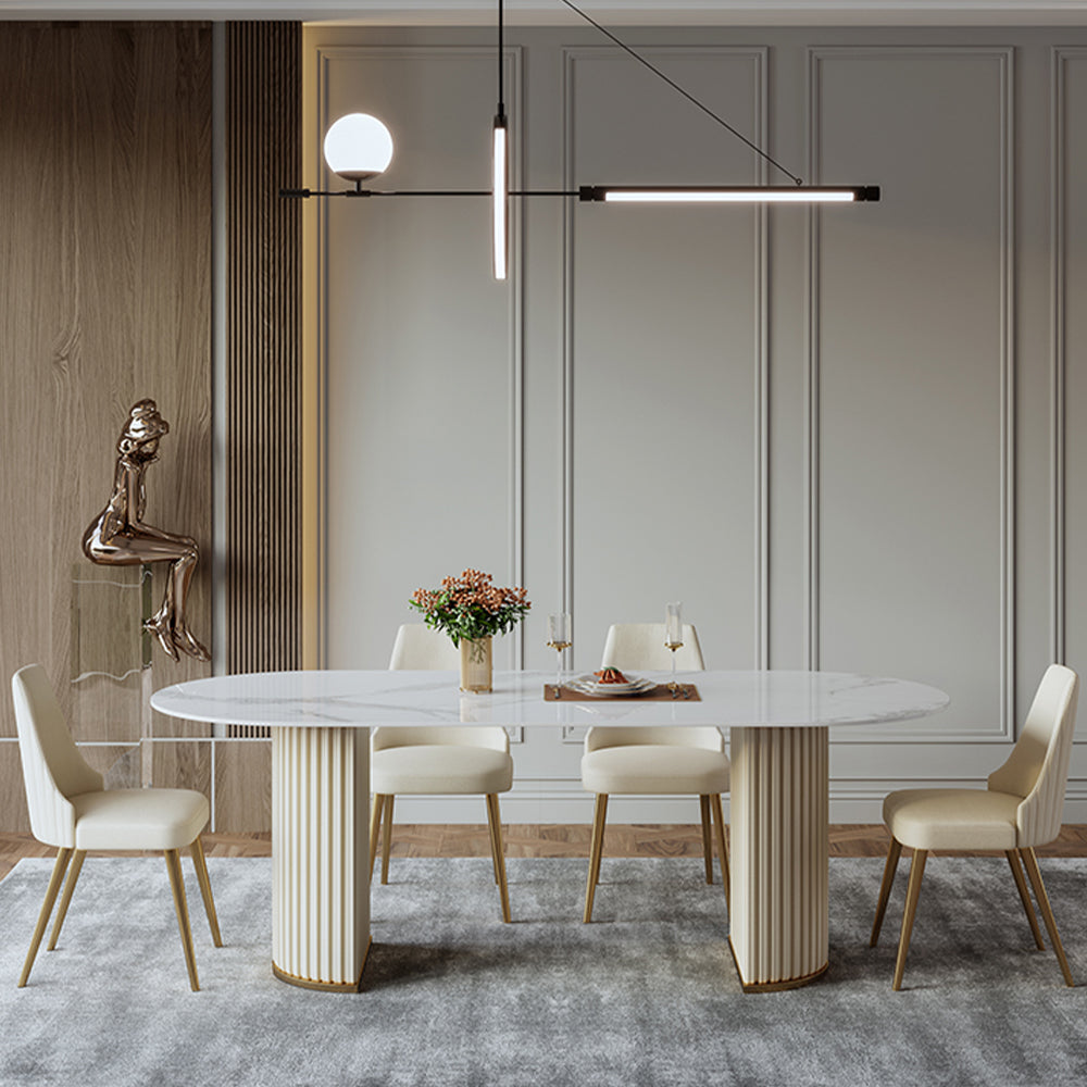 Elegant round marble dining table with fluted gold base, surrounded by cream velvet chairs on tapered gold legs, beneath a modern linear pendant light in a chic, paneled room.