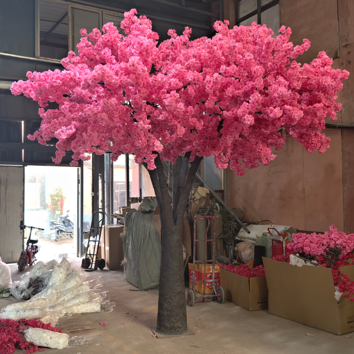 Artificial tree with striking pink blossoms, displayed in a workshop setting alongside floral materials, showcasing its readiness for rental as wedding or event decor. Are these rental trees durable for multiple setups and dismantles?