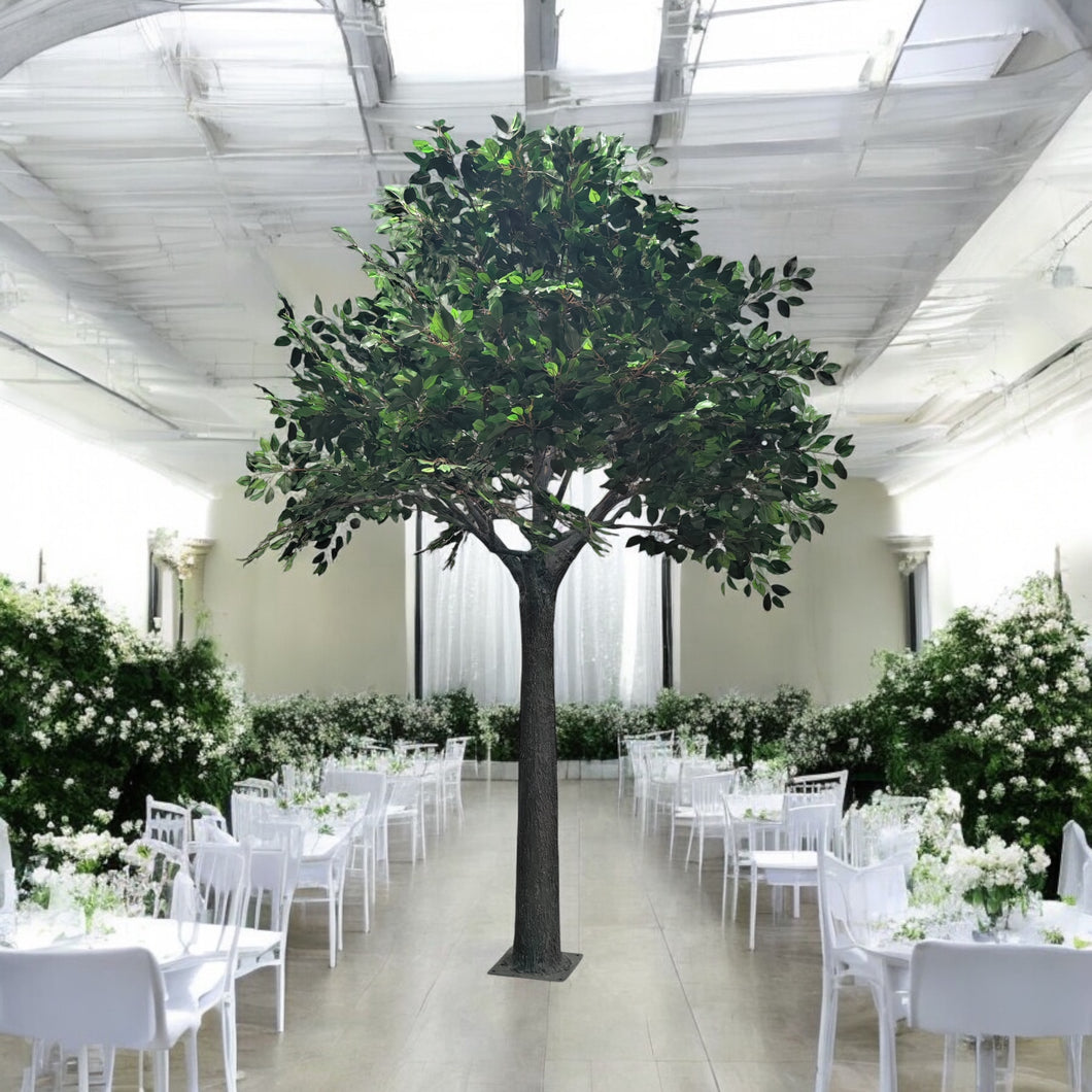 Elegant artificial green tree displayed in a beautifully decorated white event hall, surrounded by white floral arrangements and dining tables. Can this tree be used for both indoor and outdoor wedding decor?