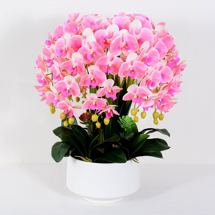A lush arrangement of artificial pink orchids with yellow buds in a simple white pot, featuring vibrant green leaves and small succulents. What are the best tips for maintaining artificial flowers to keep them looking fresh?
