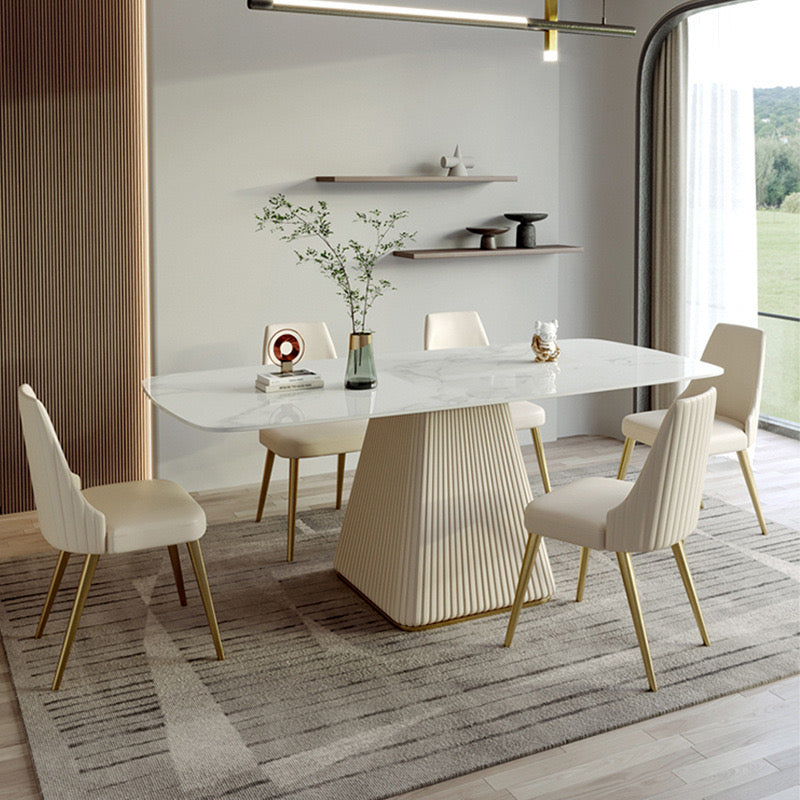 Modern rectangular marble dining table with a fluted base, paired with six cream upholstered chairs featuring gold legs, in a contemporary dining room with minimalist decor and large windows