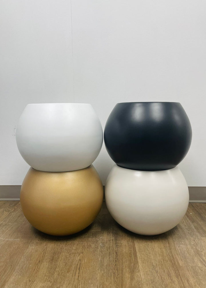 Four spherical planters in different finishes—white, black, gold, and cream—stacked in pairs, placed on a wooden floor against a plain wall.