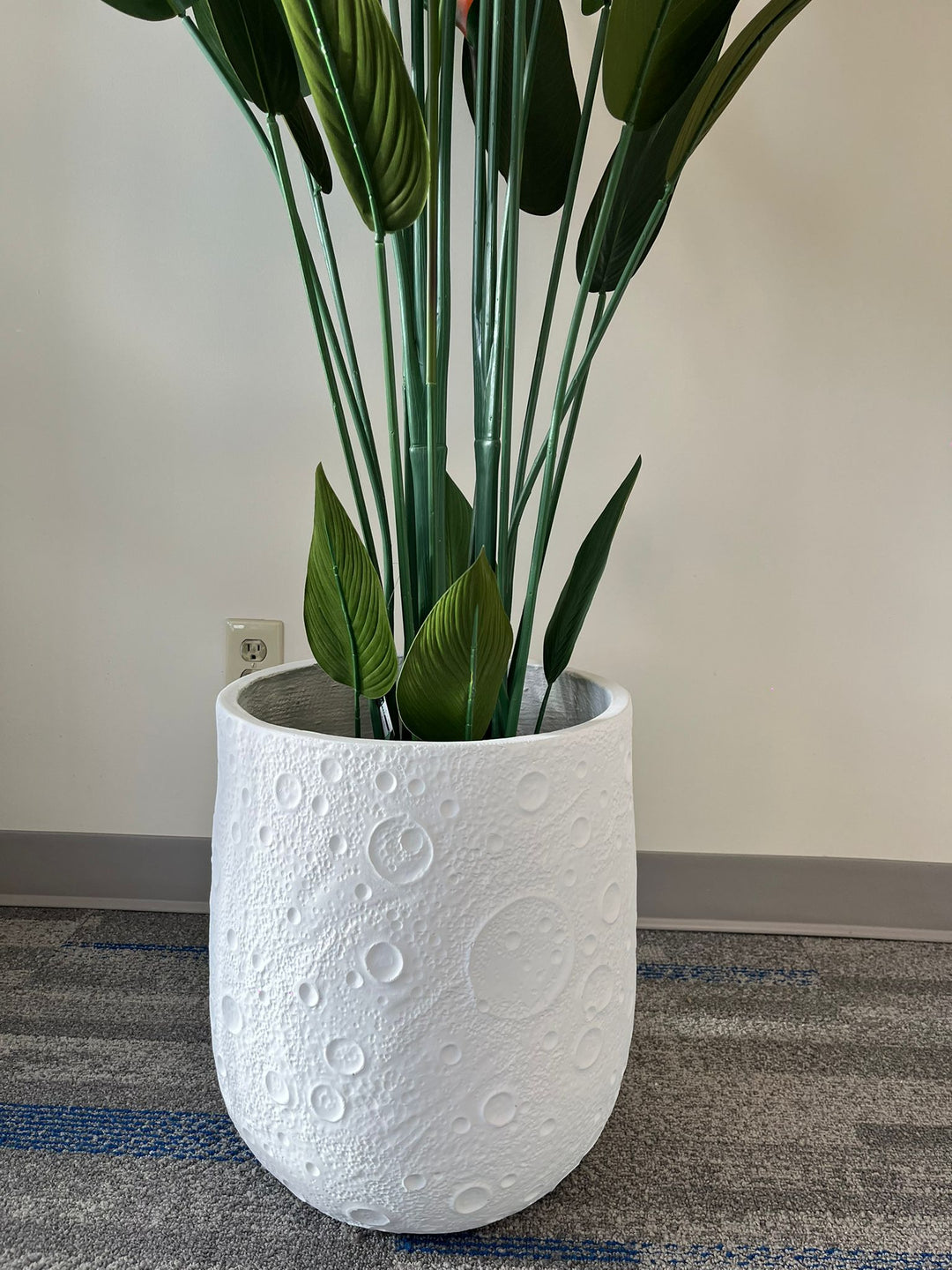 White planter with a crater-like textured design, holding an artificial bird of paradise plant with vibrant green leaves and orange flowers, placed indoors on a carpeted floor. Is this planter made from eco-friendly materials?