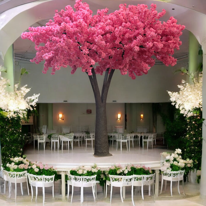 Artificial tree with vivid pink blossoms, elegantly positioned on a raised stage in a wedding venue surrounded by floral arrangements and white seating. Can these rental trees be used to create a dramatic focal point for ceremonies?