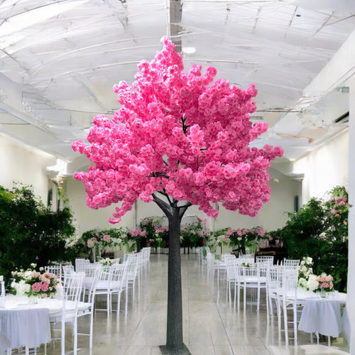 A stunning artificial pink cherry blossom tree is prominently displayed in the center of an elegant event venue. The room features white chairs and tables adorned with pastel floral arrangements, set against a backdrop of lush greenery and a high, translucent ceiling that allows soft natural light to fill the space.