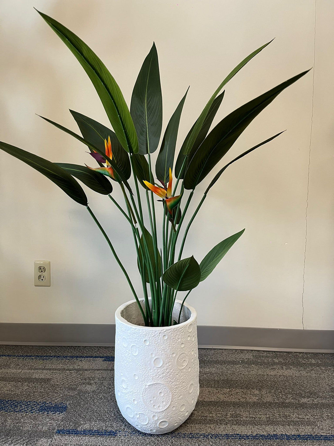 White planter with a crater-like textured design, holding an artificial bird of paradise plant with vibrant green leaves and orange flowers, placed indoors on a carpeted floor. Is this planter made from eco-friendly materials?