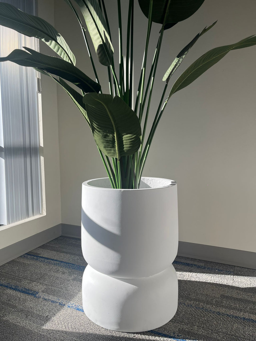 Close-up of a white stackable planter with a smooth matte finish, holding a tall artificial plant with large green leaves, placed indoors near a window with natural light. Does this planter have drainage holes?