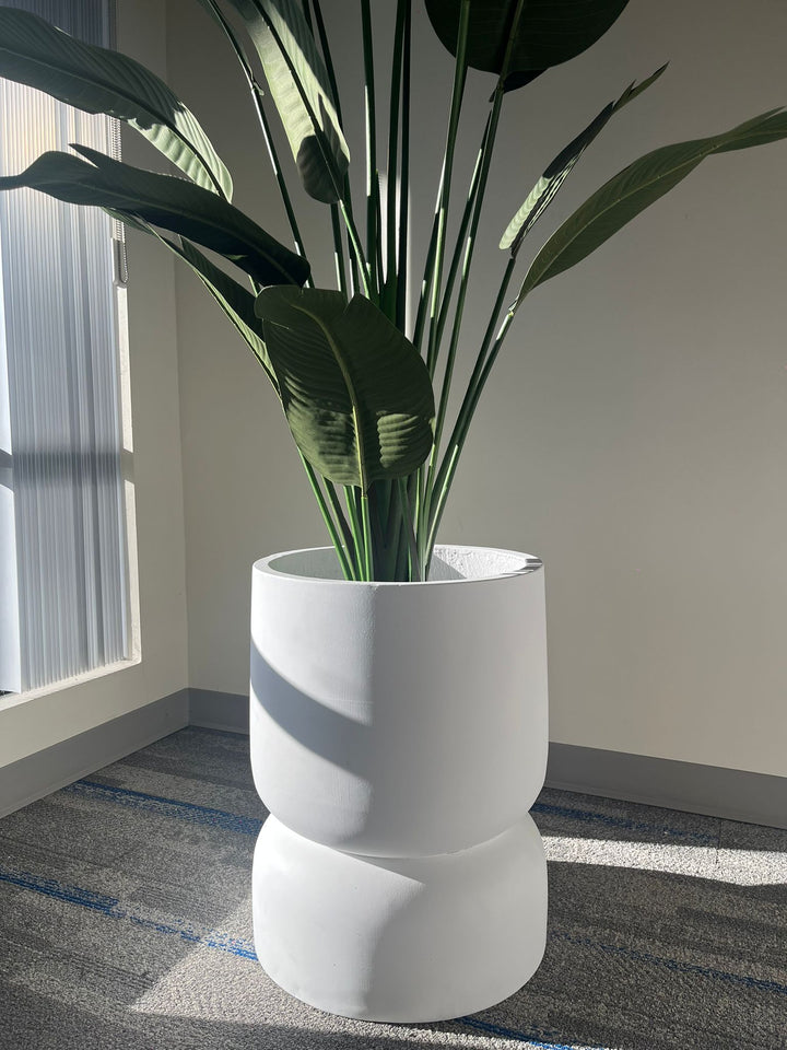 Close-up of a white stackable planter with a smooth matte finish, holding a tall artificial plant with large green leaves, placed indoors near a window with natural light. Does this planter have drainage holes?