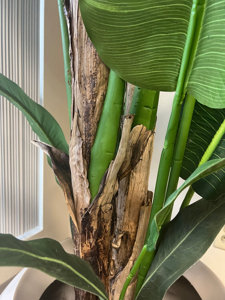 Close-up view of the base of an artificial bird of paradise plant, showing the detailed texture of the faux bark and vibrant green stems, placed in a white round planter in an office setting.