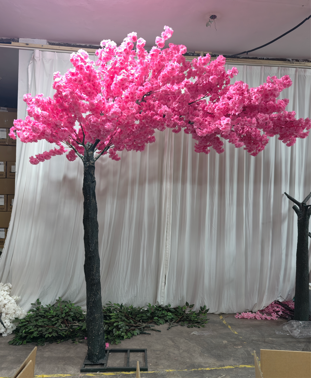 Artificial cherry blossom tree in vibrant pink, staged against a white curtain backdrop with additional floral components on the floor. Is this cherry blossom tree available for rental for indoor events?