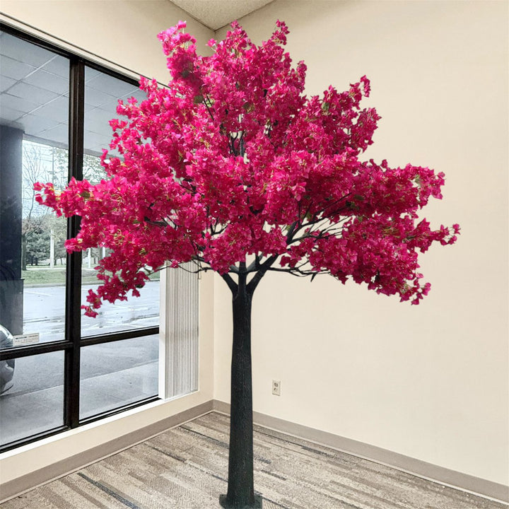 A large artificial Hot pink 10ft bougainvillea flowering tree with dense bright pink blossoms and a dark trunk, standing indoors near a window and beige wall.
