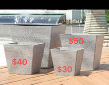 Charger l&#39;image dans la galerie, Where to buy pots. Four square-shaped outdoor planters arranged on a wooden deck. The planters feature a textured woven pattern in a light gray color. Each planter is labeled with a price in red text: $30 for the smallest, $40 for the next size, $50 for the two largest planters. The background includes water features and part of a modern architectural structure.
