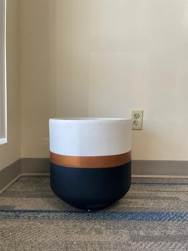 A modern, rounded planter with a matte white top, a copper stripe in the middle, and a dark navy base sits on a gray and blue carpet near a beige wall with an electrical outlet in the background.