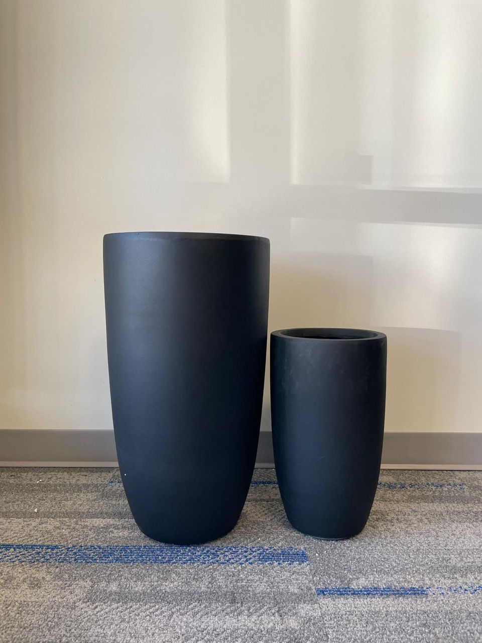 Sleek, black matte planters of different sizes. They have a minimalist, cylindrical shape with smooth surfaces and are placed on a gray carpeted floor with blue accents. The larger planter is on the left, and the smaller one is on the right. The neutral background emphasizes the modern and clean aesthetic of the planters.