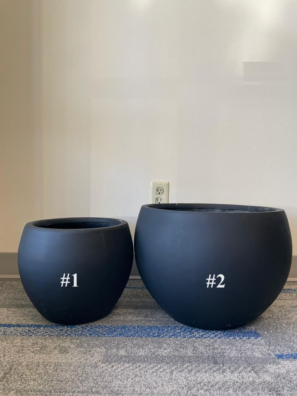 Two matte black planters of different sizes placed on a carpeted floor. The smaller planter is labeled #1, and the larger planter is labeled #2. Both planters have a round, minimalist design, perfect for modern indoor and outdoor spaces.