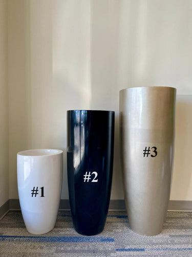 Three tall, modern planters labeled #1, #2, and #3 stand side by side on a gray and blue carpet against a light beige wall. Planter #1 is white, #2 is glossy black, and #3 is metallic gold