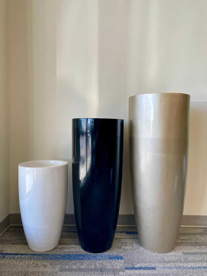 Three tall, modern planters stand side by side on a gray and blue carpet against a light beige wall. Planter #1 is white, #2 is glossy black, and #3 is metallic gold