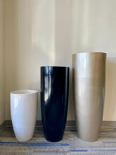 Load image into Gallery viewer, Three tall, modern planters stand side by side on a gray and blue carpet against a light beige wall. Planter #1 is white, #2 is glossy black, and #3 is metallic gold
