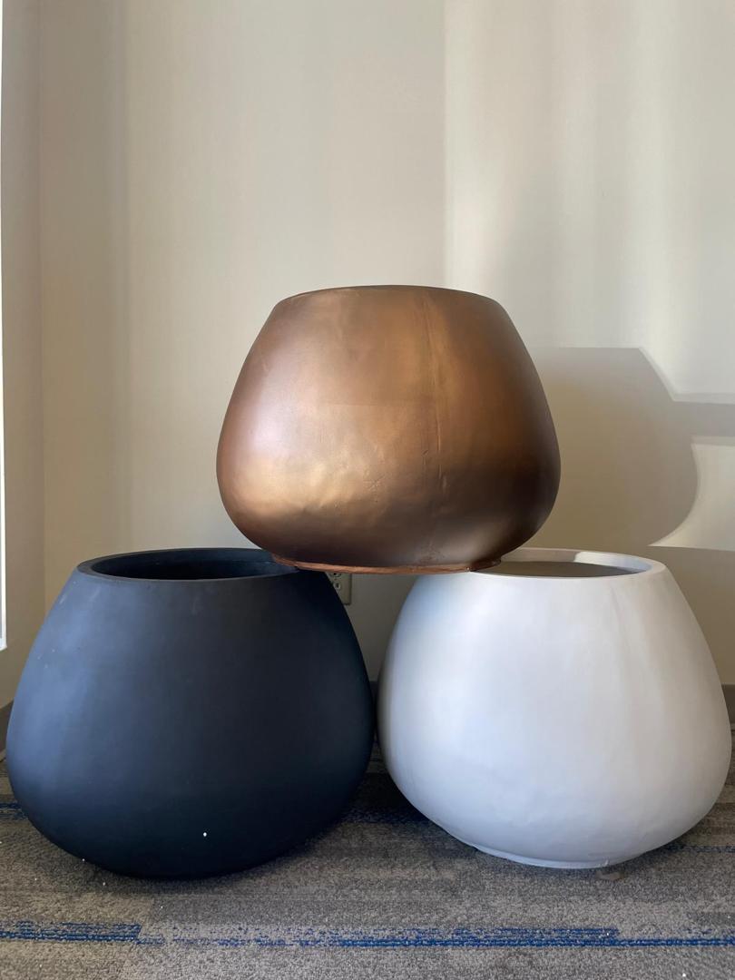 Three round planters stacked, with the top planter in a metallic bronze color and the two bottom planters in matte black and white, sitting on a carpeted floor against a light-colored wall.