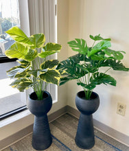 Load image into Gallery viewer, Two modern black planters with tall, ribbed bases and rounded tops, each holding lush green plants. The plant on the left has variegated leaves with yellow accents, while the plant on the right features large, vibrant green monstera leaves. The planters are placed by a window, allowing natural light to enhance the plants&#39; foliage.
