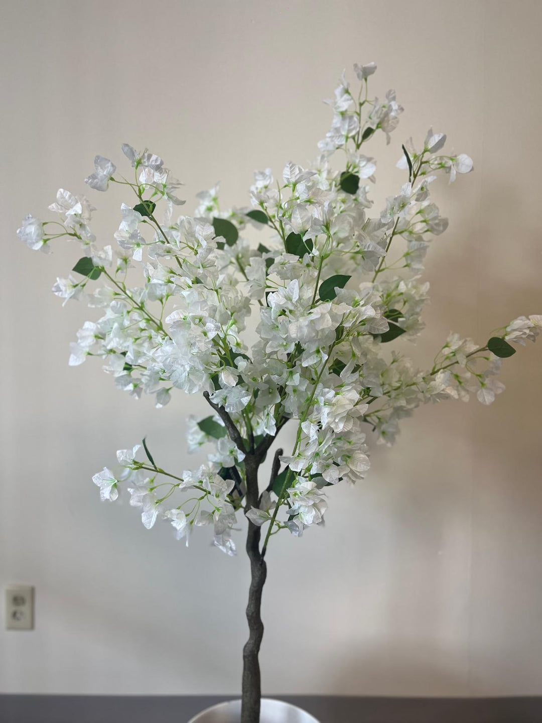 Close-up of an artificial white flower tree with multiple branches and green leaves, set against a plain beige wall.