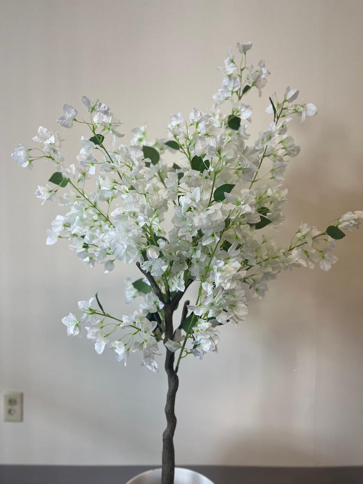 Close-up of an artificial white flower tree with multiple branches and green leaves, set against a plain beige wall.