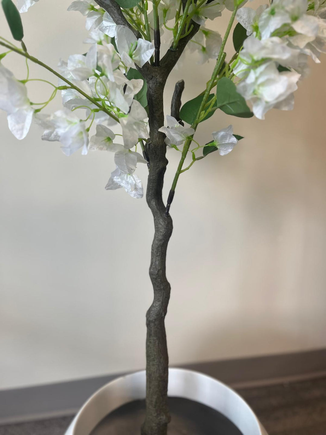Close-up of the trunk and base of an artificial white flower tree, showing the dark, textured stem placed in a white round planter