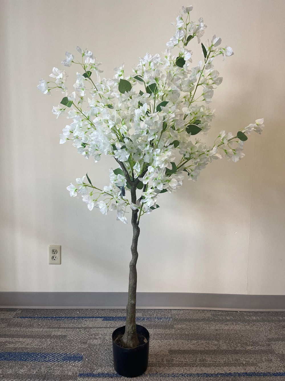 Artificial white flower tree with abundant blossoms in a modern white round planter, placed against a light-colored wall on a carpeted floor.