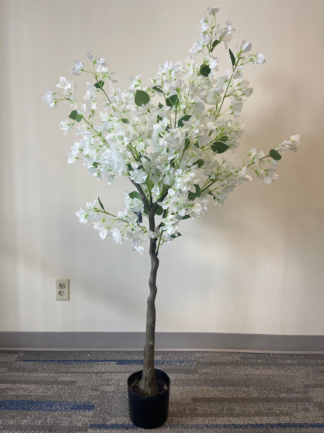 Artificial white flower tree with abundant blossoms in a modern white round planter, placed against a light-colored wall on a carpeted floor.