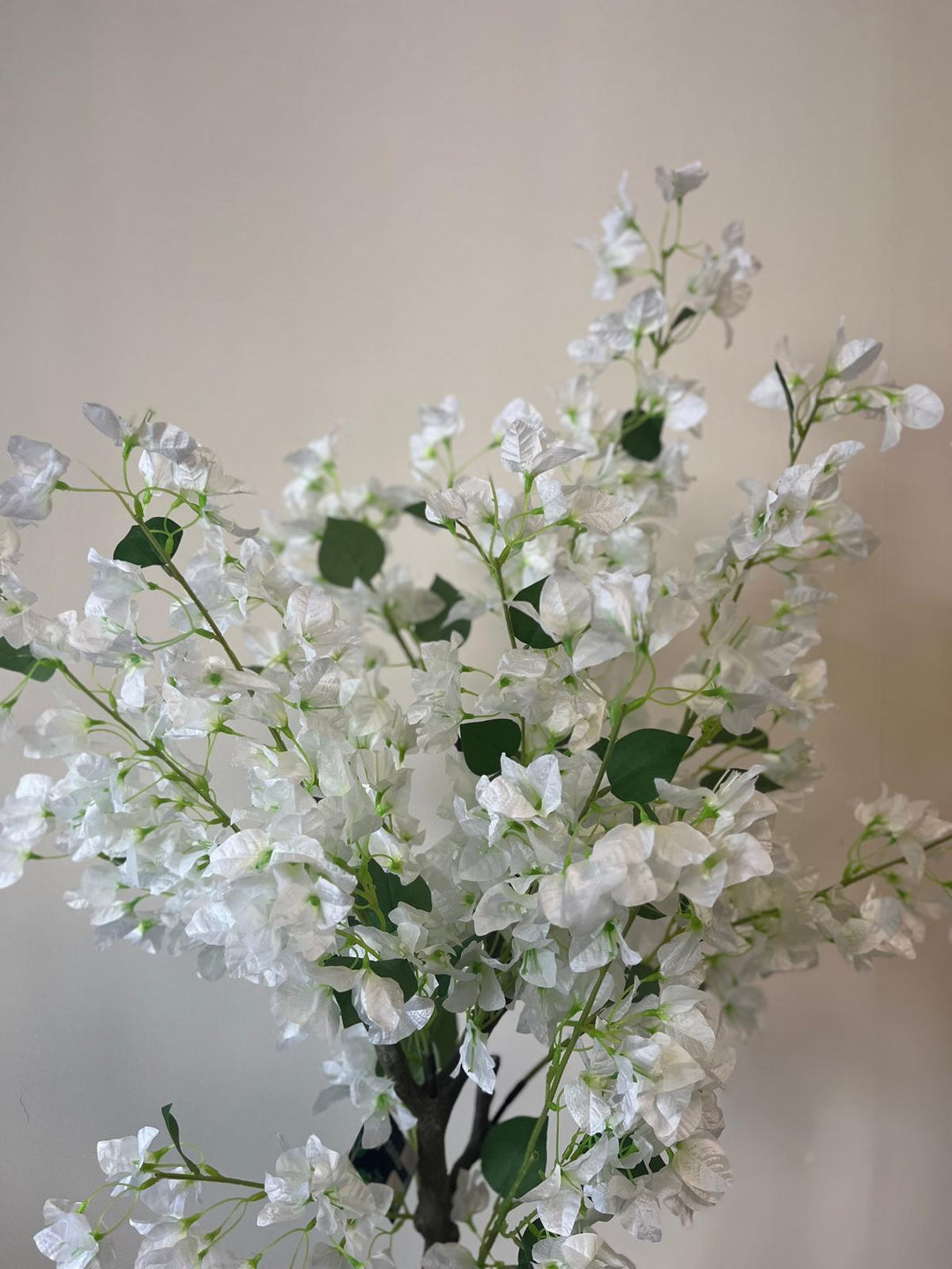 Close-up of an artificial white flower tree with delicate blossoms and green leaves, placed against a neutral-colored wall.