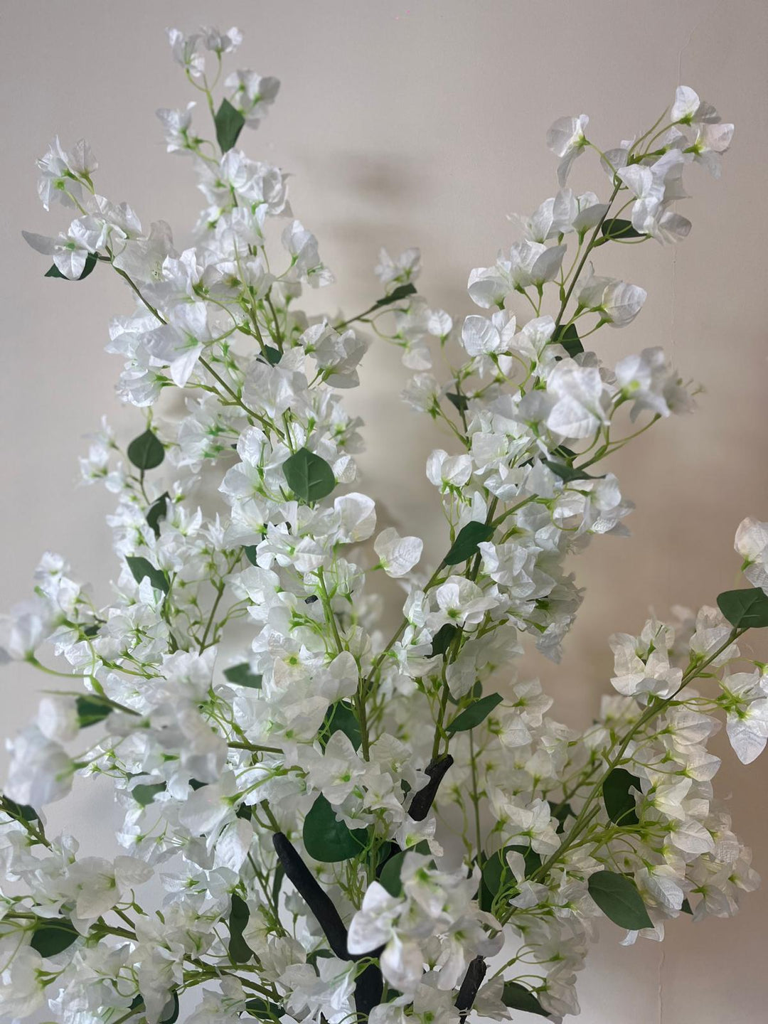 Close-up of an artificial white flower tree with delicate blossoms and green leaves, placed against a neutral-colored wall