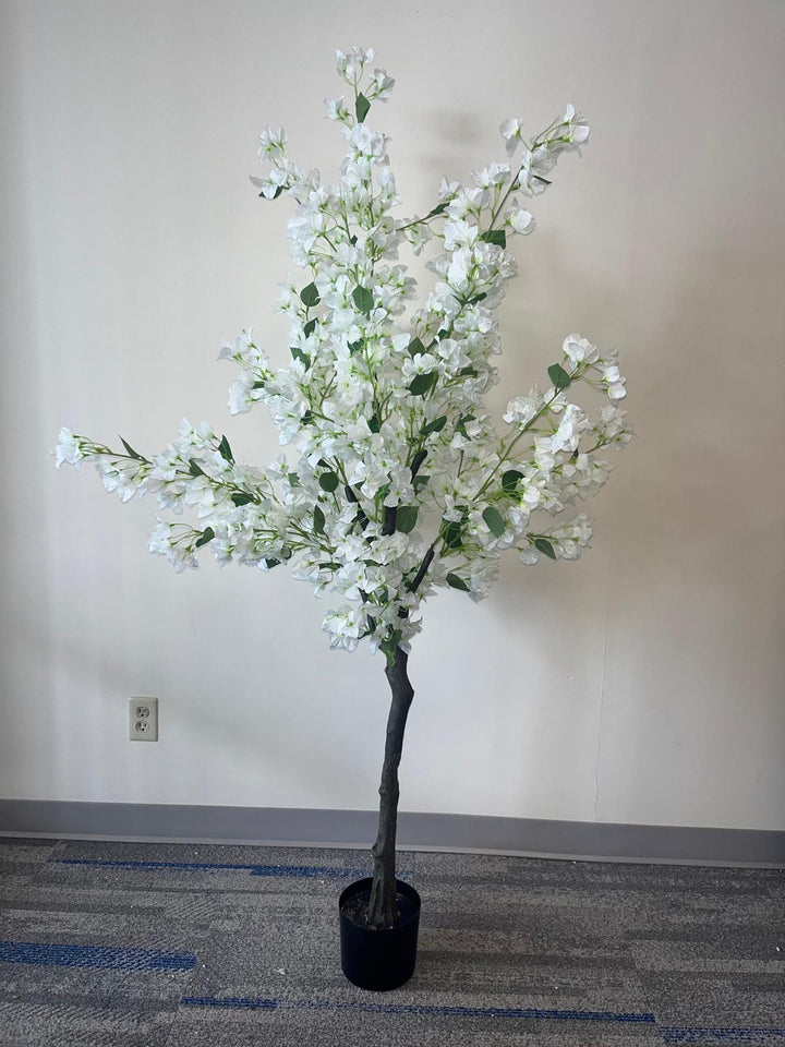 Full view of an artificial white flower tree with a dark, slender trunk, placed in a small black pot, standing against a plain wall on a carpeted floor.