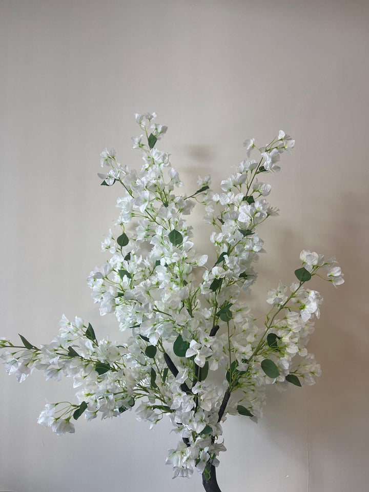Close-up of an artificial white flower tree with multiple branches and green leaves, set against a plain beige wall