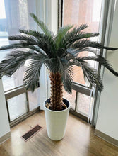 Charger l&#39;image dans la galerie, Artificial palm tree in a tall white textured planter placed in a bright corner near large windows. Common questions: What are the best artificial palm trees for indoor use? How do I choose the right planter for a large artificial tree? Can this planter be used for both real and artificial plants?
