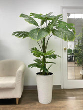 Load image into Gallery viewer, Artificial monstera plant placed in a tall, textured white planter next to a beige chair in a modern indoor setting. Commonly asked questions: What are the best artificial plants for home décor? How to choose the right planter for large artificial plants? Can monstera plants thrive indoors, and are there artificial options for a similar look?
