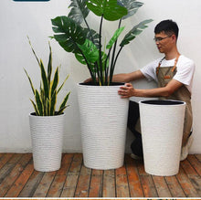 Charger l&#39;image dans la galerie, Person arranging artificial plants, including a snake plant and a monstera in tall, white textured planters of various sizes, on a wooden deck. Common questions: What is the best size planter for large artificial plants? How to choose planters for indoor vs outdoor use? Can these planters be used with real plants?
