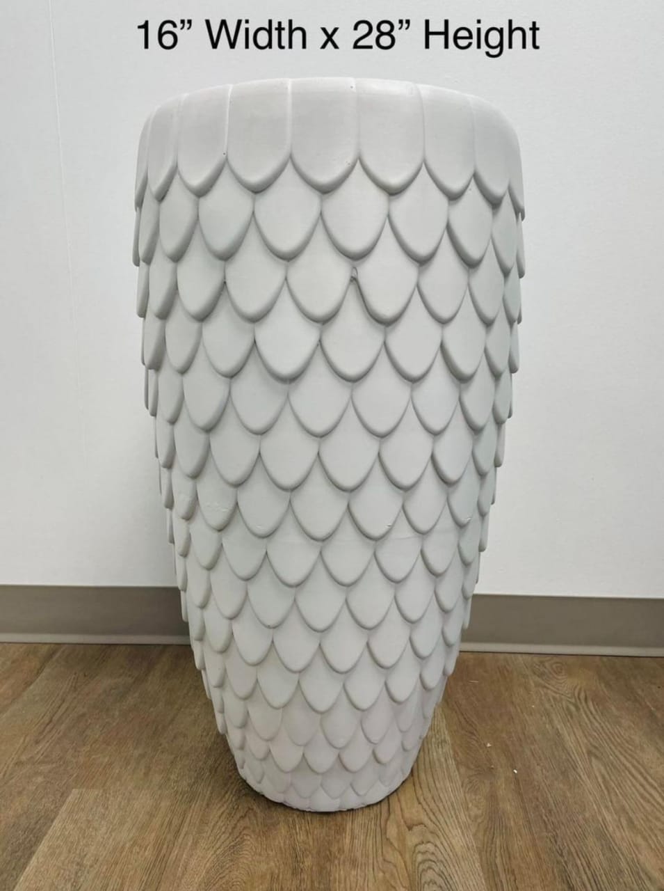 A tall white planter with a textured scale pattern, placed on a wooden floor against a neutral wall. What are the best plants to pair with textured planters for a modern home look?