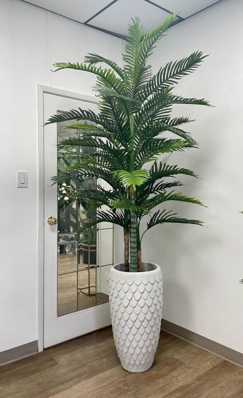 A large artificial palm plant in a white planter with a scale-like texture, placed indoors next to a door with a mirror. The plant adds a tropical touch to the modern interior setting.