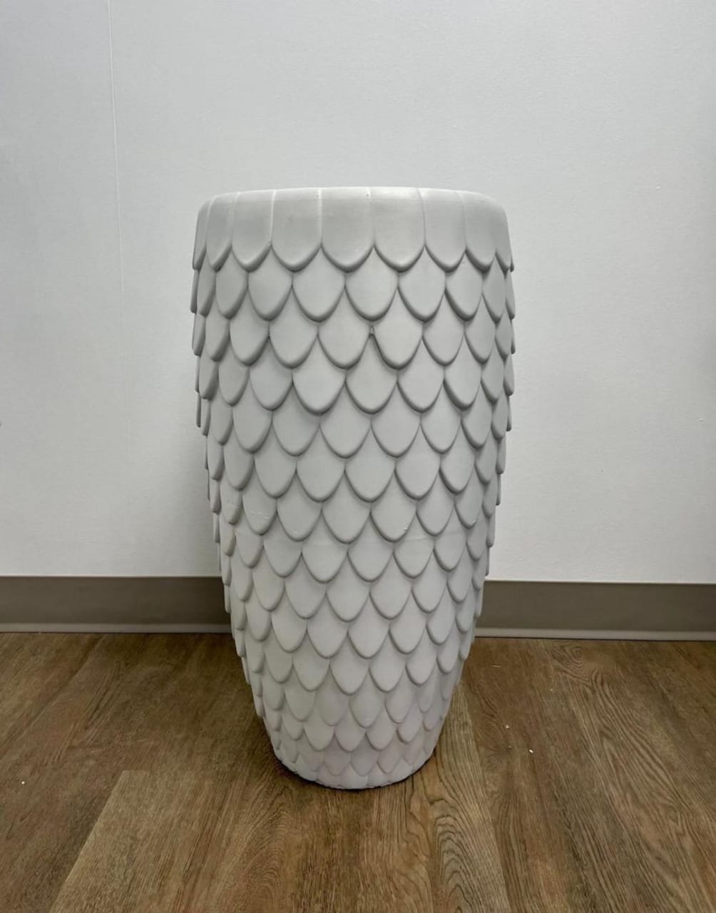 A tall white planter with a textured scale pattern, placed on a wooden floor against a neutral wall. What are the best plants to pair with textured planters for a modern home look?