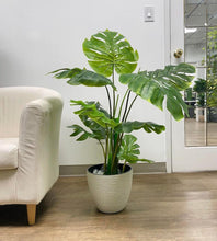 Load image into Gallery viewer, Artificial monstera plant with large, vibrant green leaves in a white textured planter, placed beside a cream-colored armchair in a modern room setting. Frequently asked questions: How realistic are artificial monstera plants? What size pot is best for indoor artificial plants? How to care for artificial plants and prevent dust build-up?
