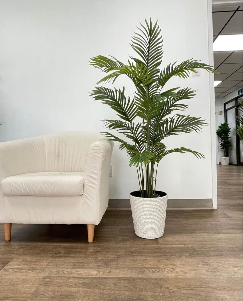 Artificial palm plant in a modern white textured planter placed beside a minimalist beige armchair on a wooden floor. Common questions: What are the benefits of artificial plants for office spaces? How to style artificial plants in living rooms? What type of planter complements minimalist decor?